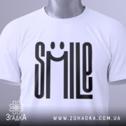 Футболка посміхнись. Біла футболка, складена на столі, фон світло-сірий, принт з написом 'SMILE' з усмішкою, на ярлику зелена стрічка.