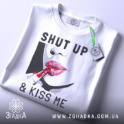 Футболка поцілуй мене, біла, зображена зверху, фон світло-сірий, принт з червоними губами та помадою, текст "SHUT UP & KISS ME", на комірці є етикетка.