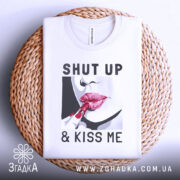 Футболка поцілуй мене, біла, зображена на плетеній кошику, фон світло-сірий, принт з червоними губами та помадою, текст "SHUT UP & KISS ME".