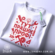 Футболка працює профі. Біла футболка, складена на поверхні, з червоним принтом, видно етикетку з зеленим шнурком, фон світло-сірий, принт містить інструменти та напис 'ТУТ ПРАЦЮЄ ПРОФІ!' у стилі графіки.