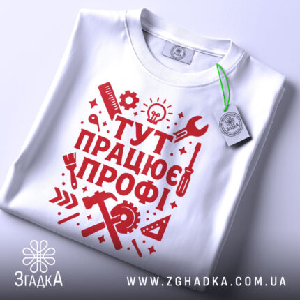 Футболка працює профі. Біла футболка, складена на поверхні, з червоним принтом, видно етикетку з зеленим шнурком, фон світло-сірий, принт містить інструменти та напис 'ТУТ ПРАЦЮЄ ПРОФІ!' у стилі графіки.