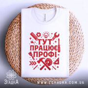 Футболка працює профі. Біла футболка, складена на плетеній кошику, з червоним принтом, фон світло-сірий, принт містить інструменти та напис 'ТУТ ПРАЦЮЄ ПРОФІ!' у стилі графіки.