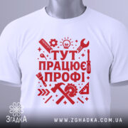 Футболка працює профі. Біла футболка, складена на поверхні, з червоним принтом, видно етикетку з зеленим шнурком, фон світло-сірий, принт містить інструменти та напис 'ТУТ ПРАЦЮЄ ПРОФІ!' у стилі графіки.