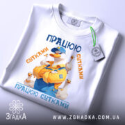 Футболка працюю сутками подарунок, біла, зображена на столі, фон світлий, принт з качкою в жовтому комбінезоні, текст 'ПРАЦЮЮ СУТКАМИ' у синіх та оранжевих кольорах, з етикеткою.