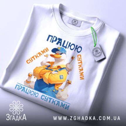 Футболка працюю сутками подарунок, біла, зображена на столі, фон світлий, принт з качкою в жовтому комбінезоні, текст 'ПРАЦЮЮ СУТКАМИ' у синіх та оранжевих кольорах, з етикеткою.