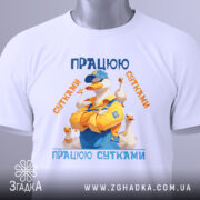 Футболка працюю сутками подарунок, біла, зображена на столі, фон світлий, принт з качкою в жовтому комбінезоні, текст 'ПРАЦЮЮ СУТКАМИ' у синіх та оранжевих кольорах, з етикеткою.