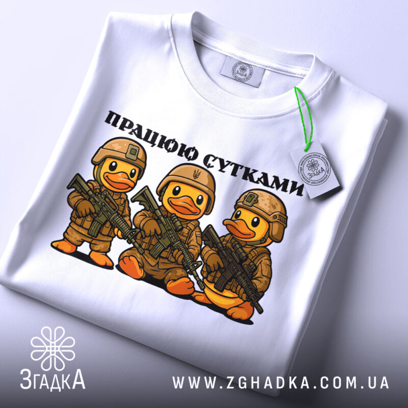 Футболка працюю сутками, біла, складена на столі, фон світлий, принт з трьома качками в камуфляжі з автоматами, текст зверху: 'ПРАЦЮЮ СУТКАМИ', на ярлику зелена стрічка.