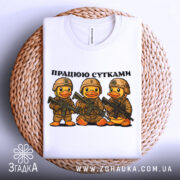 Футболка працюю сутками, біла, складена на плетеному кошику, фон нейтральний, принт з трьома качками в камуфляжі з автоматами, текст зверху: 'ПРАЦЮЮ СУТКАМИ'.