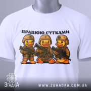 Футболка працюю сутками, біла, складена на столі, фон світлий, принт з трьома качками в камуфляжі з автоматами, текст зверху: 'ПРАЦЮЮ СУТКАМИ', на ярлику зелена стрічка.