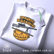 Футболка працюю віддалено, білого кольору, складена на поверхні, фон світло-сірий, принт з написом 'ПРАЦЮЮ ВІДДАЛЕНО' та зображенням гвинтівки і вимірювальної лінійки в жовтих і чорних кольорах, з етикеткою на зеленому шнурку.