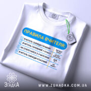 Футболка правила вчителя, біла, зображена зверху, фон нейтральний, принт з текстом правил вчителя, на футболці є етикетка з зеленим шнурком.