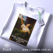 Футболка прекрасний день. Біла футболка, складена на поверхні, з принтом жінки, що тримає птаха. На футболці видно етикетку з зеленим шнурком. Принт займає центральну частину, знизу напис: 'Який прекрасний блаженний день'.