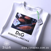 Футболка про гречку, біла, складена, фон світлий, принт з ножем, гречкою та грошима, з етикеткою на шнурку.