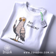 Футболка проблемний голуб, біла, складена на столі, фон світлий, принт з пташкою з ножем і текстом 'ANY PROBLEMS?', на комірці видна етикетка.