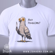Футболка проблемний голуб, біла, складена на столі, фон світлий, принт з пташкою з ножем і текстом 'ANY PROBLEMS?', на комірці видна етикетка.