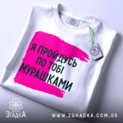 Футболка пройдусь мурашками, біла, зображена на столі, фон світлий, принт у вигляді рожевої смуги з текстом чорного кольору, на комірці є етикетка.