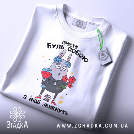 Футболка просто будь собою, білого кольору, зображена в плоскому вигляді, фон світлий, принт з малюнком зайця з квітами та келихом, текст зверху на українській мові, на комірці видно етикетку.