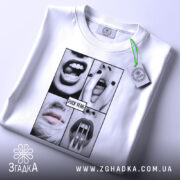 Футболка провокаційний арт, біла, складена на столі, фон світло-сірий, принт з чотирьох чорнобілих зображень з губами і зубами, текст "FUCK YEAH" на чорному фоні, на комірці видна етикетка.