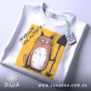 Футболка прийшов час копати. Біла футболка, складена на поверхні, з видимим принтом кота з лопатою на жовтому фоні. Принт розташований в центрі, а на комірці видно етикетку. Фон світлий, що підкреслює деталі принта.