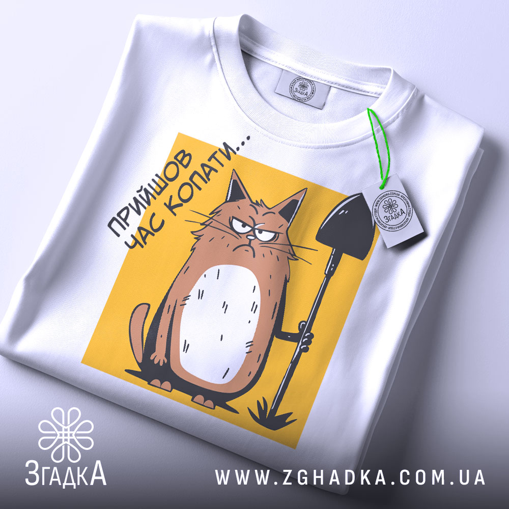 Футболка прийшов час копати. Біла футболка, складена на поверхні, з видимим принтом кота з лопатою на жовтому фоні. Принт розташований в центрі, а на комірці видно етикетку. Фон світлий, що підкреслює деталі принта.