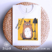 Футболка прийшов час копати. Біла футболка, складена на плетеній основі, з принтом кота з лопатою на жовтому фоні. Принт яскравий, а фон натуральний, що створює контраст і підкреслює деталі малюнка.