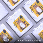 Футболка прийшов час копати. Біла футболка з принтом, на якому зображений кіт з лопатою на жовтому фоні. Кіт має сердите вираження обличчя, а принт займає центральну частину футболки. Фон нейтральний, що підкреслює кольори принта.