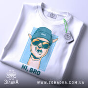 Футболка прикольна лама, біла, складена на столі, зображена з верхнього ракурсу, фон світло-сірий, принт з ламою в блакитній шапці та окулярах, текст "HI, BRO" під малюнком, з етикеткою на шнурку.