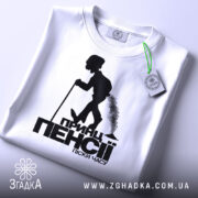 Футболка принц пенсії, біла, зображена зверху, складена на поверхні, фон світлий, принт чорного кольору з силуетом постаті, що йде з палицею, з етикеткою зеленого кольору.