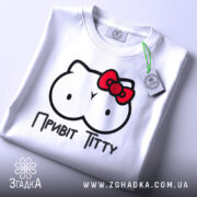 Футболка привіт ціці, біла, зображена на столі, ракурс зверху, фон світлий, принт з малюнком у вигляді двох круглих форм з очима та червоною бантиком, текст "Привіт Тітті" під малюнком, на футболці є етикетка.