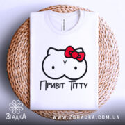 Футболка привіт ціці, біла, зображена на плоскій поверхні, фон світлий, принт з малюнком у вигляді двох круглих форм з очима та червоною бантиком, текст "Привіт Тітті" під малюнком, футболка розташована на плетеній основі.
