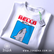 Футболка привіт від акули. Складена біла футболка з принтом акули на блакитному фоні, текст 'JUST WANTED TO SAY HELLO' червоним кольором. На футболці є етикетка з зеленим шнурком. Фон світлий, простий.