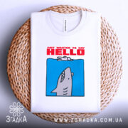 Футболка привіт від акули. Складена біла футболка з принтом акули на блакитному фоні, текст 'JUST WANTED TO SAY HELLO' червоним кольором. Футболка розташована на плетеній основі. Фон світлий, простий.