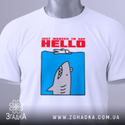 Футболка привіт від акули. Складена біла футболка з принтом акули на блакитному фоні, текст 'JUST WANTED TO SAY HELLO' червоним кольором. На футболці є етикетка з зеленим шнурком. Фон світлий, простий.
