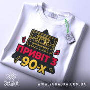 Футболка привіт з 90-х, біла, складена на поверхні, фон світло-сірий, принт з касетою та написом 'ПРИВІТ З 90-х' у червоному та жовтому кольорах, з етикеткою на комірці.