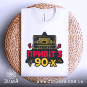 Футболка привіт з 90-х, біла, складена на плетеній кошику, фон світло-сірий, принт з касетою та написом 'ПРИВІТ З 90-х' у червоному та жовтому кольорах.