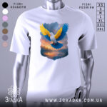 Футболка птах свободи, біла, зображена на манекені, фон світло-сірий, принт з птахом з жовтими та блакитними крилами на фоні заходу сонця.