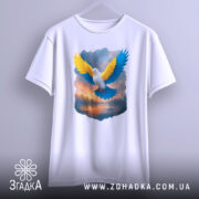 Футболка птах свободи, біла, підвішена на вішалці, фон світло-сірий, принт з птахом з жовтими та блакитними крилами на фоні заходу сонця.