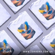 Футболка птах свободи, біла, зображена на манекені, фон світло-сірий, принт з птахом з жовтими та блакитними крилами на фоні заходу сонця.