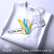 Футболка птах волі білого кольору, складена на столі, ракурс зверху, фон світло-сірий, принт з птахом у жовтих та блакитних тонах, напис 'птах волі' на стрічці, з етикеткою зеленого кольору.