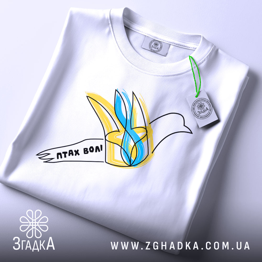 Футболка птах волі білого кольору, складена на столі, ракурс зверху, фон світло-сірий, принт з птахом у жовтих та блакитних тонах, напис 'птах волі' на стрічці, з етикеткою зеленого кольору.