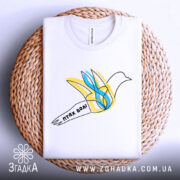 Футболка птах волі білого кольору, складена на плетеному кошику, ракурс зверху, фон світло-сірий, принт з птахом у жовтих та блакитних тонах, напис 'птах волі' на стрічці.