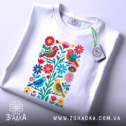 Футболка пташки у квітах, біла, складена на поверхні, фон світло-сірий, принт з пташками і квітами, з біркою на комірці.