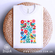 Футболка пташки у квітах, біла, складена на плетеній кошику, фон світло-сірий, принт з пташками і квітами, видно етикетку на комірці.