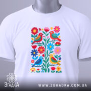 Футболка пташки у квітах, біла, складена на поверхні, фон світло-сірий, принт з пташками і квітами, з біркою на комірці.