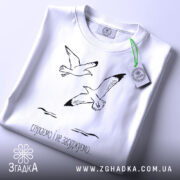 Футболка пташина свобода, білого кольору, зображена зверху, на столі, фон світло-блакитний, принт з двома птахами та текстом "слухаємо і не засуджуємо...", на футболці є етикетка з зеленим шнурком.