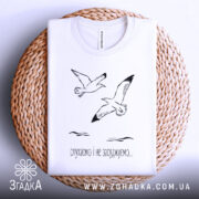 Футболка пташина свобода, білого кольору, складена на плетеній кошику, фон світло-блакитний, принт з двома птахами та текстом "слухаємо і не засуджуємо...".