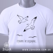 Футболка пташина свобода, білого кольору, зображена зверху, на столі, фон світло-блакитний, принт з двома птахами та текстом "слухаємо і не засуджуємо...", на футболці є етикетка з зеленим шнурком.