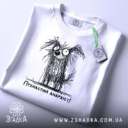 Футболка пухнастий анархіст, білого кольору, складена на плоскій поверхні, фон світло-сірий, принт з малюнком пухнастого собаки з великими очима та написом 'Пухнастий анархіст!' під ним, з етикеткою на зеленому шнурку.