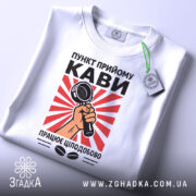 Футболка пункт прийому кави, біла, складена на столі, фон світлий, принт з червоними променями та мікрофоном у руці, на комірці зелена етикетка.