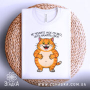Футболка пузіко нажріть своє, біла футболка, зображена зверху на плетеній кошику, фон світло-сірий, принт з котом, що усміхається, текст українською мовою.
